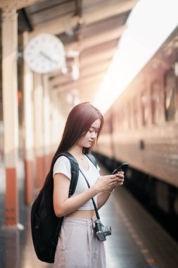 Sırt çantası ve kamerası olan genç bir kadın tren istasyonunda beklerken akıllı telefonunu kullanıyor, yumuşak güneş ışığıyla yıkanmış..