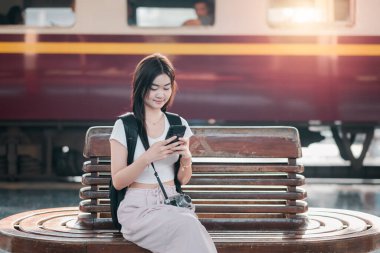 Bir tren istasyonunda ahşap bir bankta oturan genç bir kadın, arka planda antika bir trenle akıllı telefonunu kullanıyor..