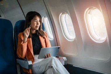 Profesyonel bir kadın uçakta tablet kullanıyor. Pencereden güneş ışığının ve manzaranın tadını çıkarıyor. İş seyahatini ve teknolojiyi temsil ediyor..