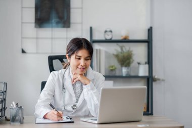 Modern bir ofiste dizüstü bilgisayarla çalışan beyaz önlüklü kadın bir doktor kalemle not alıyor..