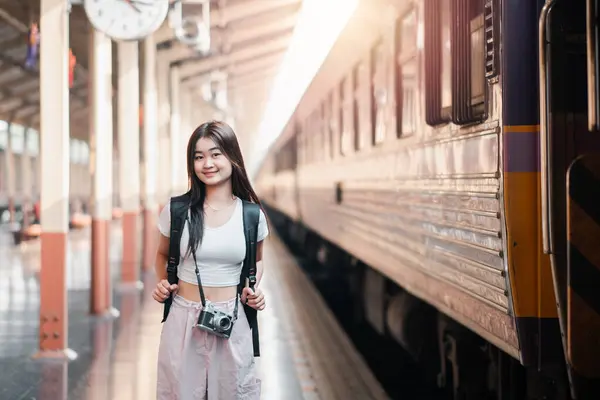 stock image A young woman with a camera and backpack stands on a train station platform, ready for travel. Sunlight filters through the station.