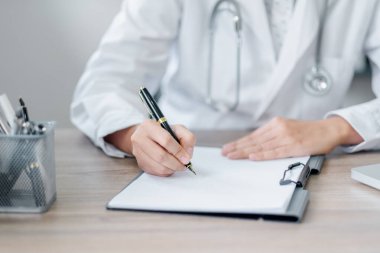 Sağlık hizmetleri ve profesyonelliği vurgulayan, sağlık ofisindeki bir masanın panosuna yazı yazan beyaz önlüklü bir doktorun yakın plan görüntüsü..