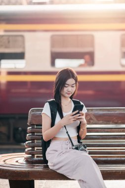 Tren istasyonunda bir bankta oturan genç bir kadın, akıllı telefonunu kullanıyor. Sırt çantası ve kamerası var. Seyahat etmeyi ya da işe gitmeyi öneriyor..