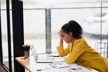 Modern bir ofis masasında dizüstü bilgisayarda çalışan sarı ceketli genç bir kadın odaklanmış ve düşünceli görünüyor..