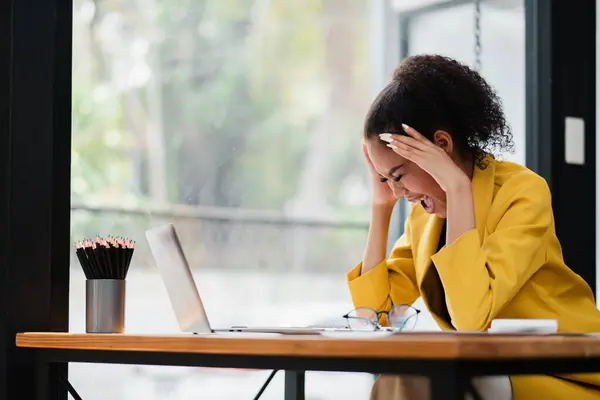 stock image A stressed businesswoman in a yellow blazer sits at her office desk, holding her head in frustration while working on her laptop.