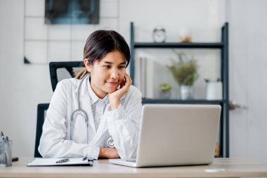 Beyaz önlüklü genç bir bayan doktor modern ofis ortamında dizüstü bilgisayarda çalışıyor. Arka planda tıbbi ekipmanlar ve kitaplık var..