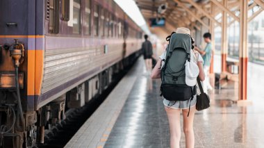 Büyük bir sırt çantasıyla bir tren platformu boyunca yürüyen maceraya hazır bir gezgin. Tren istasyonunun arka planında bir tren ve diğer yolcular var..