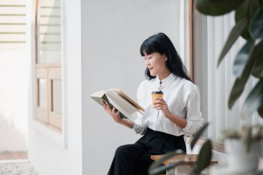 Genç bir kadın, bir kitap okuyup, modern minimalist bir ortamda kahvesini yudumlayarak huzurlu bir anın tadını çıkarıyor..