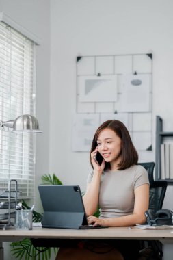 Evde çalışan, tablet kullanan ve telefonla konuşan, modern ofis ekipmanları ve doğal ışık ile çevrili genç bir kadın..