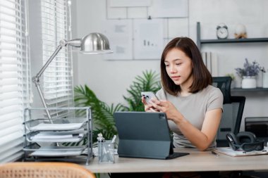 Modern ofis malzemeleri ve dekor ile çevrili akıllı telefon ve tablet kullanarak ev ofisinde birden fazla iş yapan genç bir kadın..