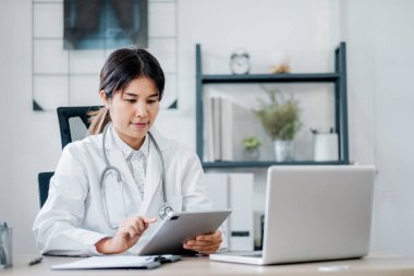 Modern ofis ortamında dijital tablet kullanan beyaz önlüklü kadın doktor dizüstü bilgisayar ve tıbbi ekipman kullanıyor..