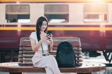 Tren istasyonunda bir bankta oturan genç bir kadın, akıllı telefonunu kullanıyor. Sırt çantası ve arka planda tren olan bir kamerası var..