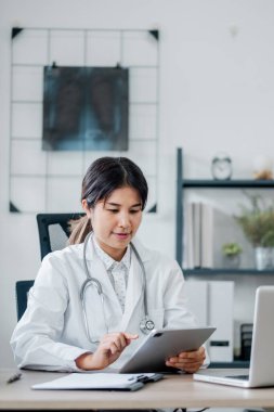 Beyaz önlüklü kadın doktor modern bir muayenehanede dijital tablet kullanıyor ve arka planda röntgen görüntüsü var..