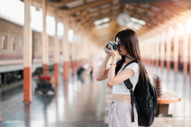 Sırt çantası ve antika kameralı genç bir kadın tren istasyonunda fotoğraf çekiyor. Seyahat ve maceranın özünü yakalıyor..