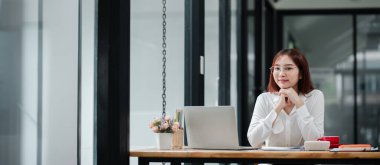 Beyaz gömlekli profesyonel bir kadın, cam duvarlı, modern bir ofiste, ahşap bir masada dizüstü bilgisayarla çalışıyor..