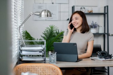 Ev ofisinden çalışan genç bir kadın telefonla konuşurken gülümsüyor ve tablet, modern ve düzenli çalışma alanı kullanıyor..
