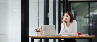 Beyaz gömlekli genç bir iş kadını, cam duvarları ve doğal ışığı olan modern bir ofiste ahşap bir masada dizüstü bilgisayarla çalışıyor..