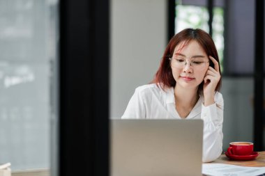 Modern ofis ortamında dizüstü bilgisayarda çalışan beyaz gömlekli genç bir kadın. Masasında kırmızı bir kahve fincanı var..
