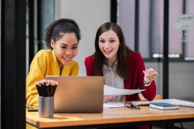 Modern bir ofiste bir projede birlikte çalışan iki profesyonel kadın dizüstü bilgisayara bakarken gülümseyip fikirleri tartışıyorlar..