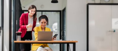 Modern bir ofiste birlikte çalışan iki profesyonel kadın dizüstü bilgisayar ve akıllı telefon kullanarak takım çalışması ve üretkenlik sergiliyorlar..