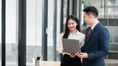 Resmi giyinmiş iki iş adamı, geniş pencereli, modern bir ofis ortamında dizüstü bilgisayarda iş tartışıyorlar..