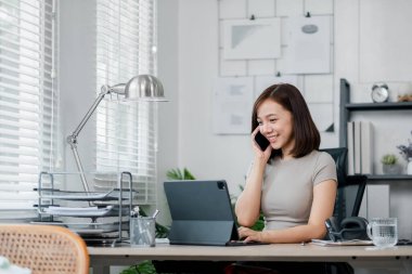 Gülümseyen genç bir kadın modern bir ofis ortamında tablet ve telefonla birden fazla iş yapıyor..