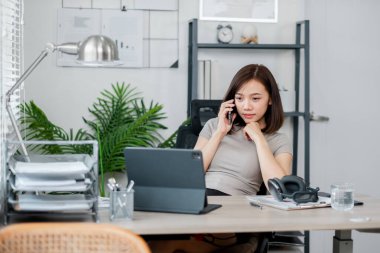 Ofiste çalışan, telefonla konuşan, dizüstü bilgisayar kullanan ve belgeleri inceleyen genç bir kadın. Fabrikalar ve ofis malzemeleriyle modern çalışma alanı.