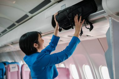 Flight attendant in blue uniform placing a bag in the overhead compartment inside an airplane cabin. clipart