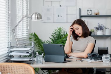 İç işlerinde çalışan genç bir kadın, dizüstü bilgisayarla çalışırken stresli görünüyor. Fabrikalar, raflar ve ofis malzemeleriyle modern çalışma alanı.