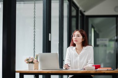 Gözlüklü genç bir kadın dizüstü bilgisayar ve belgelerle modern ofis ortamında çalışıyor..