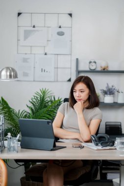 Ofis masasında çalışan genç bir kadın arka planda belgeler ve bitkilerle dizüstü bilgisayarına odaklanıyor..