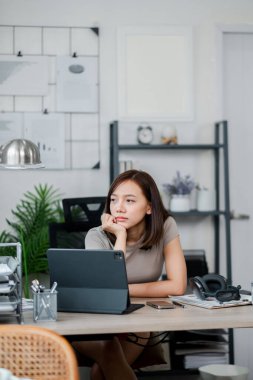 Genç bir kadın modern bir ev ofisinin masasında oturmuş, kulaklıkları yanında, çağdaş dekorlarla çevrili bir tablet üzerinde çalışıyor..