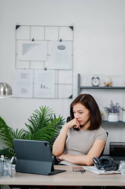 Modern bir ofis ortamında tablet üzerinde çalışan genç bir kadın. Etrafı doküman ve dekorla çevrili bitkiler ve raflarla dolu..