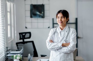 Steteskoplu beyaz önlüklü kendine güvenen bir kadın doktor modern bir muayenehanede düşünceli bir şekilde duruyor..