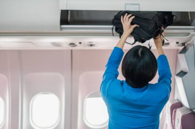 Flight attendant in blue uniform placing a black bag in the overhead compartment of an airplane cabin. clipart