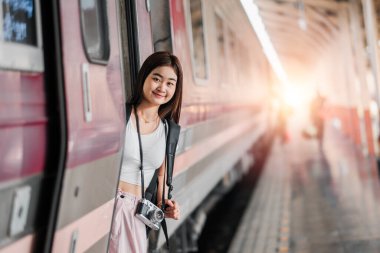 Elinde kamera olan genç bir kadın tren istasyonundaki platformdan aşağı eğiliyor. Arka planda güneş batarken gülümsüyor..