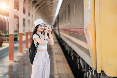 Sırt çantalı ve şapkalı genç bir kadın tren istasyonunda eski bir kamerayla fotoğraf çekiyor..