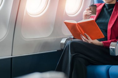 Kırmızı ceketli bir iş kadını uçuş sırasında bir uçakta oturmuş kitap okuyor ve kahve içiyor..