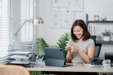 Evdeki ofis ortamında akıllı telefonunu kullanırken gülümseyen genç bir kadın. Çalışma alanı bir dizüstü bilgisayar, belgeler ve modern dekor içerir..