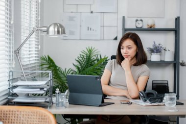 Modern ev ofisinden uzaktan çalışan genç bir kadın dizüstü bilgisayar kullanıyor ve etrafı evrak ve ofis malzemeleriyle çevrili..