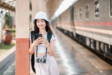 Kameralı ve akıllı telefonlu genç bir kadın gezgin tren istasyonunda duruyor, bir sonraki macerasına hazır..