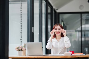 Genç profesyonel bir kadın, modern bir ofis masasında bilgisayarla çalışırken gözlüklerini ayarlıyor ve üretken bir çalışma ortamı yaratıyor..