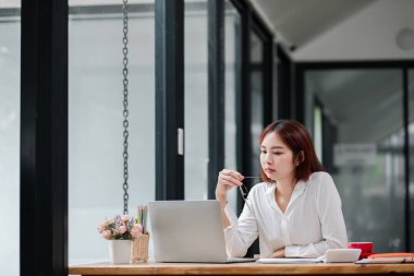 Beyaz gömlekli, odaklanmış genç bir kadın geniş pencereleri ve doğal ışığı olan, modern, parlak bir ofiste dizüstü bilgisayarla çalışıyor..