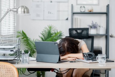 Modern bir ev ofisinde, dijital cihazlar ve ofis malzemeleriyle çevrili bir masada dinlenmekten yorgun düşmüş bir kadın..