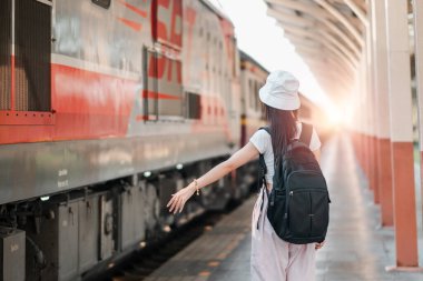 Güneş doğarken tren istasyonunda şapka ve sırt çantası takan genç bir kadın, seyahat ve macerayı simgeliyor..