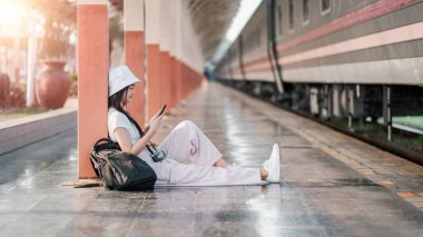 Genç bir kadın akıllı telefonunu kullanarak tren peronunda oturuyor. Beyaz şapka, sırt çantası ve kamera takıyor..