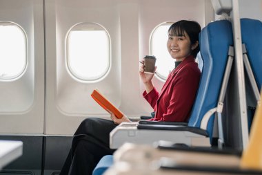 Kırmızı ceketli bir iş kadını uçakta otururken kahve içip kitap okuyor ve uçuş konforu ve verimliliğini gösteriyor..
