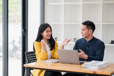 Modern bir ofiste tartışan iki genç profesyonel, ahşap bir masada belgelerle birlikte bir dizüstü bilgisayar üzerinde çalışıyorlar..