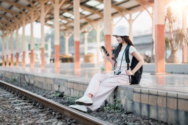 Akıllı telefonuyla tren peronunda oturan genç bir kadın. Sırt çantası ve kamera ile gündelik giyinmiş. Seyahat ve macerayı temsil ediyor..