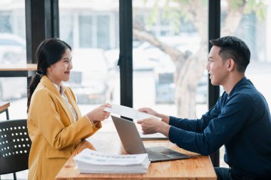 İki profesyonel iş görüşmesinde, modern bir ofiste dokümanları doğal ışıkla değiş tokuş ediyorlar..